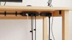 Under-desk mounted power strip and cable hub placed at rear center with neatly routed cords and a single exit path down the desk leg to the wall outlet